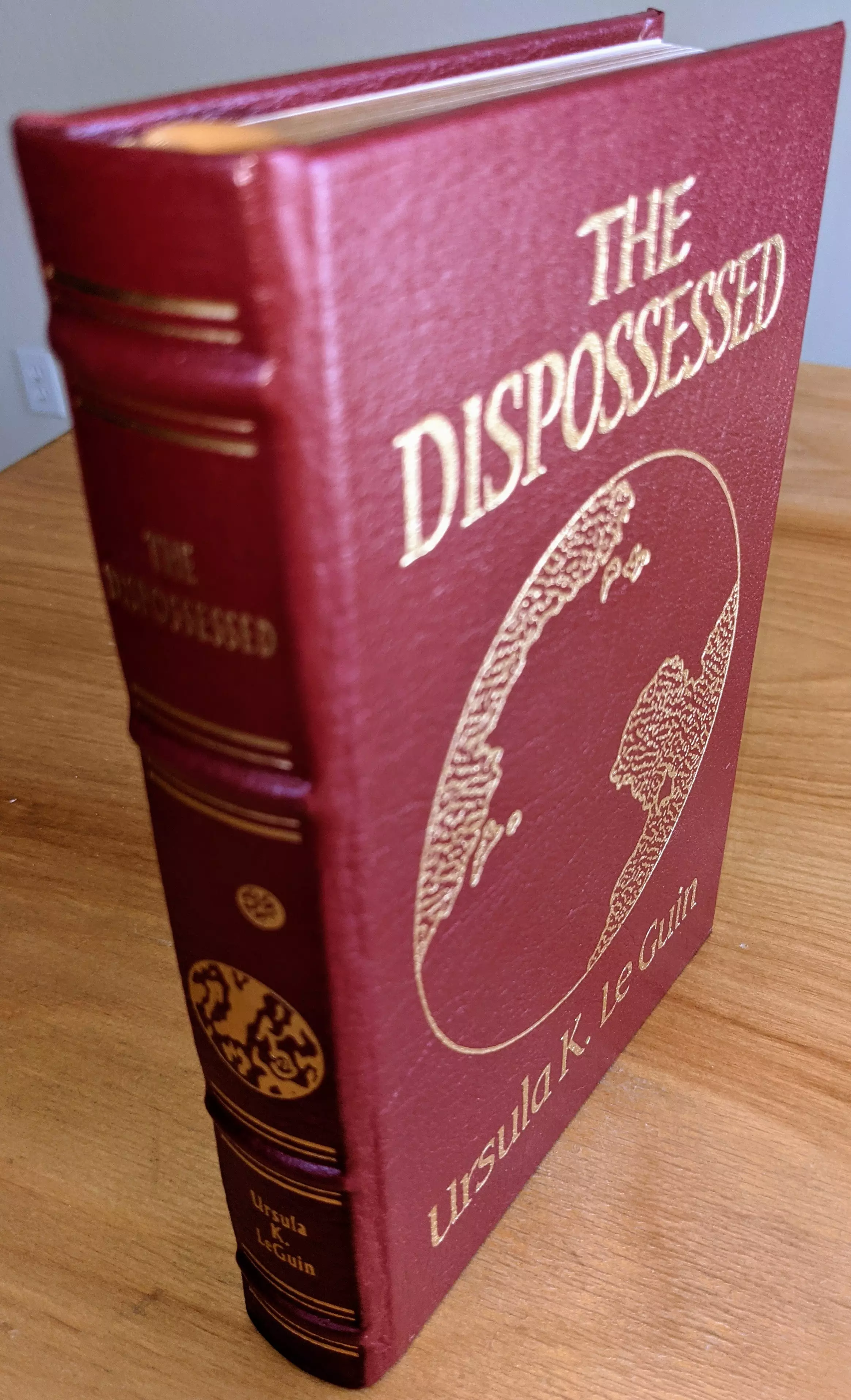 This is a pristine, leather-bound Collector’s Edition of The Dispossessed by Ursula K. Le Guin, published by The Easton Press in 1986 as part of the Masterpieces of Science Fiction Collection. Includes a special introduction by Frederik Pohl. Bound in wine colored leather with hubbed spines, the book is accented in 22kt gold.
 -
The front cover prominently features the title The Dispossessed in bold, capitalized gold lettering above an intricately detailed image of a planetary map, beautifully rendered in 22kt gold. Beneath the illustration, the author’s name, Ursula K. Le Guin, is elegantly embossed in gold.
 - 
The spine features the title and the author’s name in gold serif font, accompanied by decorative gold accents and a circular celestial motif, all divided by thin gold lines that add to the spine’s refined aesthetic.
 - 
The pages are printed on archival-quality, acid-neutral paper, ensuring longevity and preservation. The page edges are gilded, creating a radiant gold shimmer when viewed from the side. Inside, the book includes a pristine bookplate and features an original frontispiece artwork by Pat Morressey. The smyth-sewn binding ensures durability, while concealed muslin joints provide additional structural integrity.
 - 
Meticulously preserved in a dust-free bookcase within a dry climate by a single private owner, this volume remains in pristine condition.
 - 
A stunning and rare collectible for science fiction enthusiasts and fine book collectors alike.