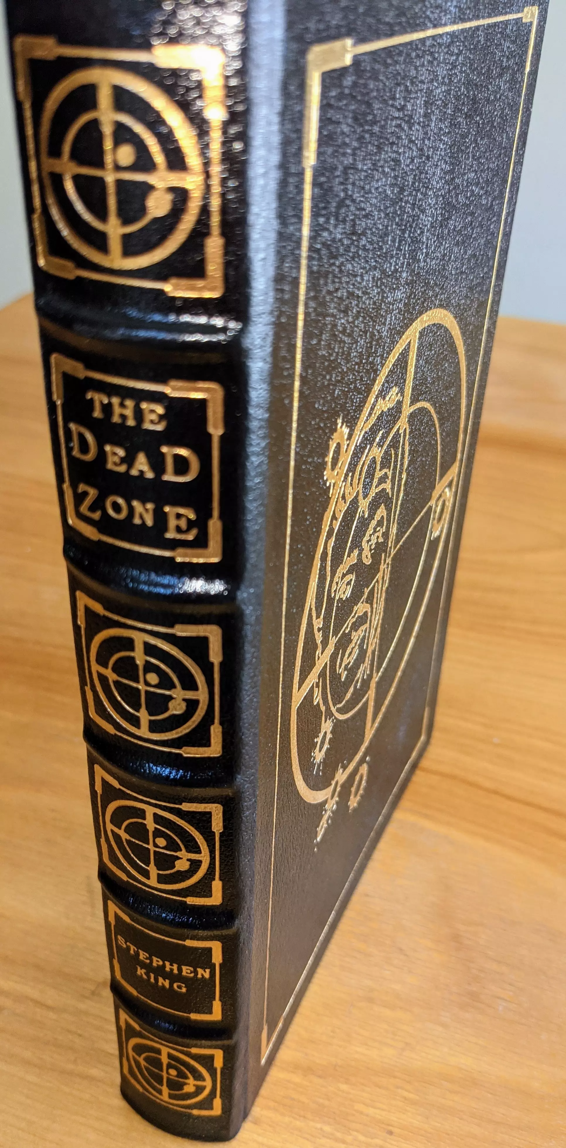 Pristine, leather-bound Collector Edition of The Dead Zone by Stephen King, published by The Easton Press in 1993 as part of the Masterpieces of Science Fiction Collection. Introduction by James Gunn
 - 
Beautifully bound in black leather, with elegant 22k gold-foil embossing. The cover and spine are adorned with intricate 22k gold-embossed designs that perfectly complement the dark and suspenseful themes. The title The Dead Zone is prominently displayed on the front cover and spine in bold gold serif lettering. Surrounding the text is an intricate design of concentric circles and crosshairs, evoking the novel's themes of fate and foresight. The cover also features an etched portrait surrounded by cosmic and geometric motifs, creating a visual connection to the narrative.
 - 
The pages are printed on archival-quality, acid-neutral paper. The page edges are 22k gold gilded. Inside, the book includes a pristine bookplate and features an original frontispiece artwork by Jill Bauman. The smyth-sewn binding ensures durability, while concealed muslin joints provide additional structural integrity
 - 
Meticulously preserved in a dust-free bookcase within a dry climate by a single private owner, this volume remains in pristine condition
 - 
A stunning and rare collectible for science fiction enthusiasts and fine book collectors alike