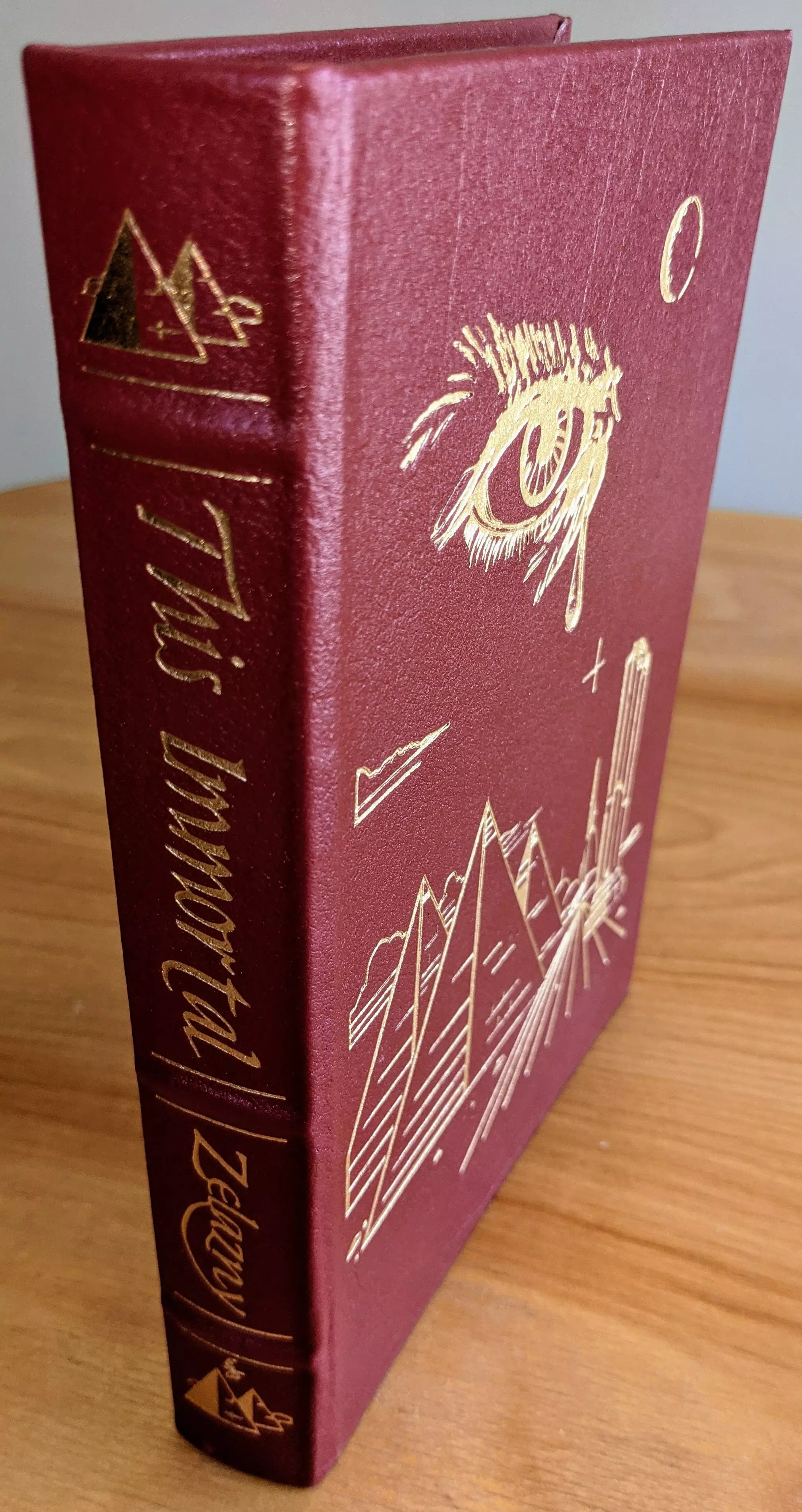 Striking maroon leather-bound edition of This Immortal by Roger Zelazny, part of The Masterpieces of Science Fiction Collection by The Easton Press. The cover and spine are intricately adorned with elegant gold-embossed designs. The title is prominently displayed on the spine in graceful gold lettering, alongside the author's name. The spine also features a small, detailed depiction of pyramids
 - 
On the cover is a bold illustration of pyramids, a glowing celestial eye, and cosmic elements, all rendered in shimmering 22kt gold
 - 
This collector's edition is expertly crafted in full leather with raised hubbed spines, ensuring durability and a classic aesthetic. It is printed on archival-quality acid-neutral paper with gilded edges, combining elegance with longevity. The binding features smyth-sewn construction and concealed muslin joints, offering both durability and ease of handling
 - 
Maintained in pristine condition, this volume is part of a single-owner private collection, stored unread in dust-free bookcases within a dry climate. A pristine, untouched bookplate is included on the inside front cover, signifying its exclusivity. This edition features an introduction by Robert Silverberg and stunning illustrations by Vincent DiFate, capturing the imaginative essence of Zelazny's storytelling. This volume is a masterpiece of literary artistry and craftsmanship, making it a prized addition for collectors
