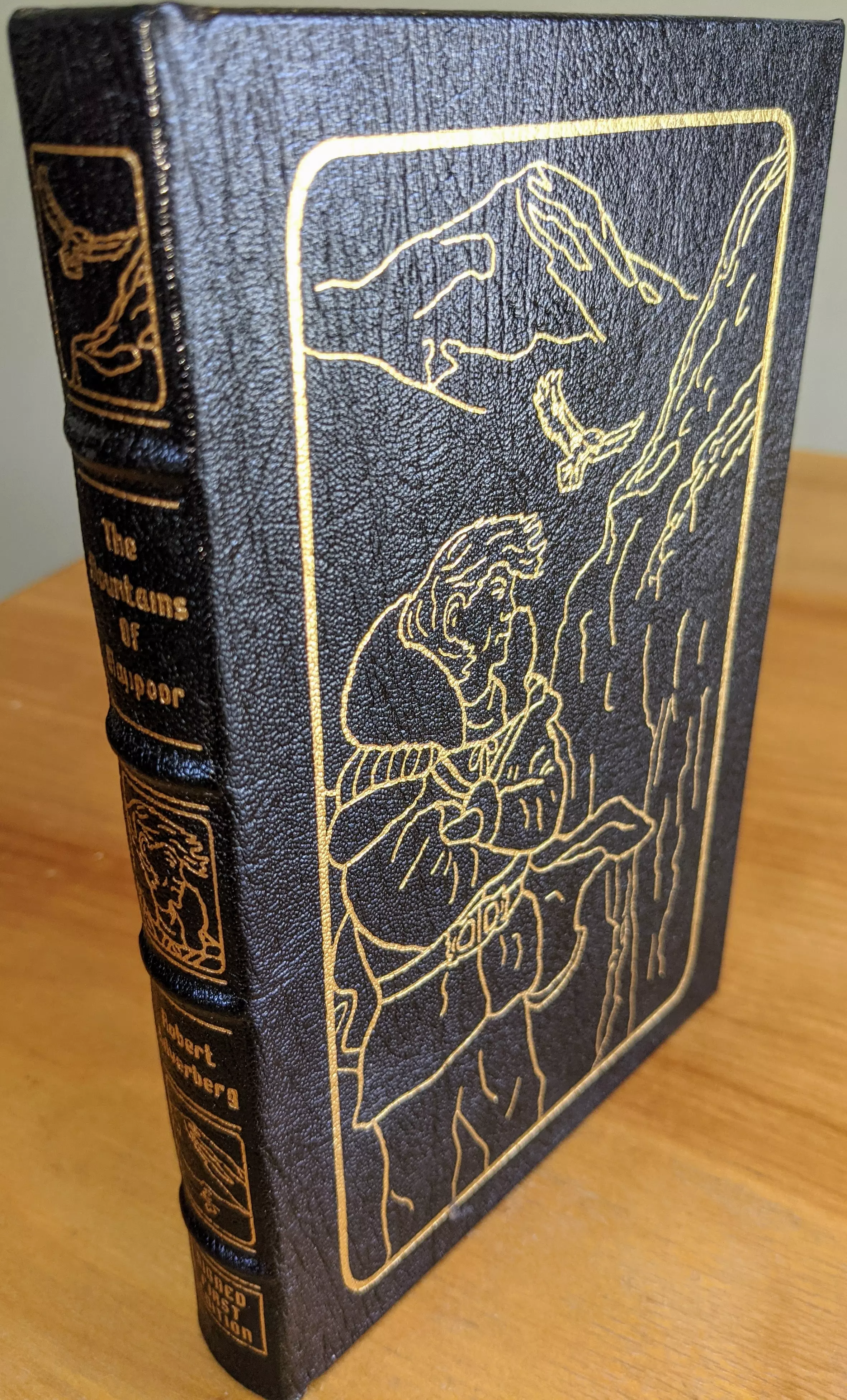 Stunning, SIGNED, (1995) FIRST Edition, black leather-bound book titled The Mountains of Majipoor by Robert Silverberg, part of The Signed First Editions of Science Fiction Collection by The Easton Press. The spine and cover are accented with 22k gold-embossed details. The spine lists the title and author's name in an elegant serif font, complemented by a mystical creature design at the bottom
 - 
The front cover features a gold-embossed illustration of a man in a fantastical setting, looking towards a towering cliff with ethereal beings, aligning with the book's otherworldly theme
 - 
This collector's SIGNED, FIRST edition is bound in full leather with raised hubbed spines, accented in 22kt gold, and printed on archival-quality acid-neutral paper with 22k gold gilded edges. It includes smyth-sewn binding and concealed muslin joints, ensuring durability and ease of handling
 - 
Stored unread in a dust-free bookcase in a dry climate, the book is in pristine condition within a private single-owner collection. It includes a pristine bookplate inside the front cover, is SIGNED by the author, and features an introduction by Jame Gunn with illustrations by Ron Miller. This makes it a prized collector's item for science fiction fans