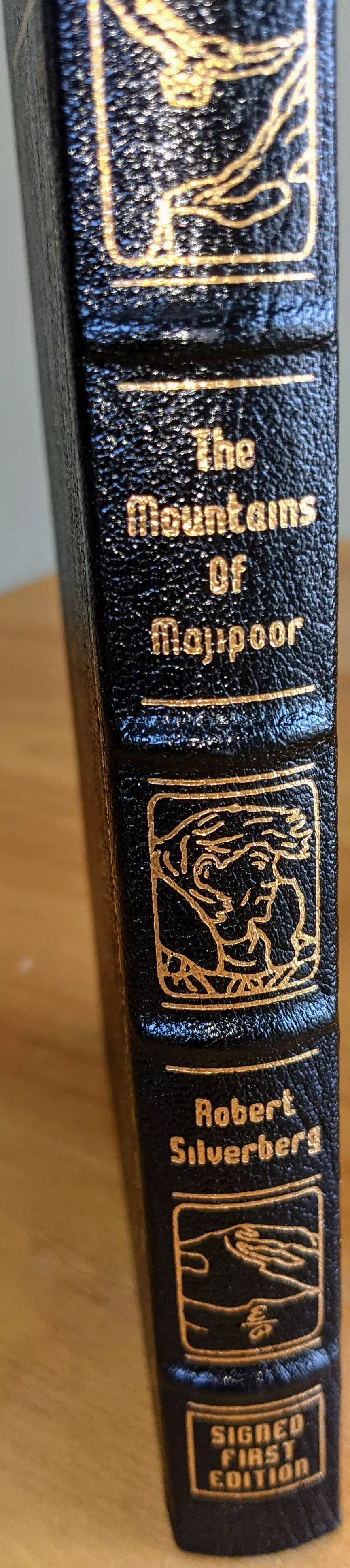 Luxurious signed first edition of The Mountains of Majipoor by Robert Silverberg, published by The Easton Press. The book is bound in full black leather, giving it a distinguished and elegant appearance. The spine is adorned with 22kt gold accents, featuring the book's title, author's name, and intricate gold-embossed artwork. The spine showcases three distinct gold-stamped illustrations: at the top, a hand extending toward another; in the middle, a detailed portrait of a man with wavy hair, possibly a character from the novel; and at the bottom, two hands clasping, reinforcing a theme of connection or unity. Below this final illustration, a gold-stamped plaque designates it as a signed first edition, further enhancing its collectible value. The hubbed spine and smyth-sewn binding ensure durability, while the archival-quality pages with gilded edges add to its luxurious feel. This exquisite edition is a fine example of Easton Press craftsmanship, making it a prized possession for collectors of classic science fiction literature.
