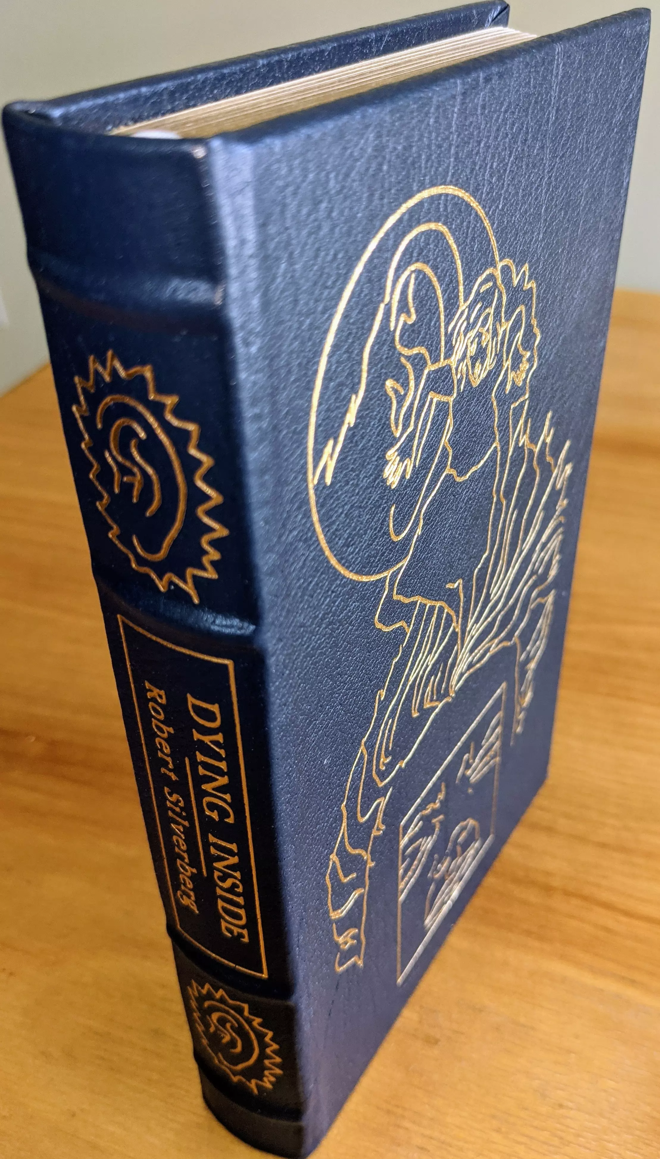 Stunning navy blue leather-bound edition of Dying Inside by Robert Silverberg, published by The Easton Press (1991). The front cover features a striking depiction of a human figure surrounded by abstract, swirling patterns, rendered in shimmering 22kt gold. These intricate designs are complemented by geometric panels
 - 
The spine displays the title Dying Inside in bold gold serif font, along with the author's name, Robert Silverberg. The design includes a stylized brain motif encircled by radiant lines. The full leather binding with raised hubbed spines ensures durability and timeless appeal
 - 
This edition is printed on archival-quality acid-neutral paper with 22k gold gilded edges, combining visual brilliance with preservation. Its smyth-sewn binding and concealed muslin joints provide structural integrity and ease of handling. Maintained in pristine, unread condition, the book has been stored in dust-free bookcases within a dry climate as part of a single-owner private collection. A pristine, untouched bookplate is included inside the front cover
 - 
This edition also features an introduction by Richard D. Erlich, and includes stunning illustrations by Frank Kelly Freas. This volume is a testament to both literary and artistic excellence, making it a prized addition to any collection