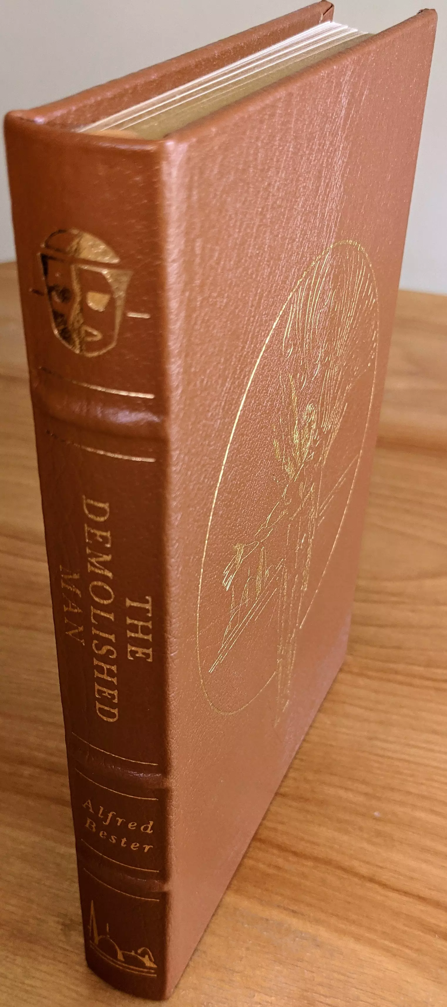 This is the Easton Press collector's edition of The Demolished Man by Alfred Bester, issued as part of the Masterpieces of Science Fiction collection. The book is bound in full tan leather with hubbed spines and accented in 22kt gold. The spine features four raised bands and gold-stamped elements, including the title THE DEMOLISHED MAN, the author's name Alfred Bester, and two decorative motifs: a stylized mask icon near the top and a minimalist cityscape at the bottom
 - 
The front cover displays original artwork by Vincent Di Fate, embossed in gold. The illustration depicts a stylized human figure in motion, overlaid by a radiant circular field. The design is geometric and abstract, evoking a sense of dynamic energy and psychological complexity, consistent with the novel's themes. Clean lines and spatial balance define the aesthetic of the composition, framed within a large circular outline
 - 
This limited-run collector's edition includes an introduction by Kurt Vonnegut. It is printed on archival-quality paper with gilded page edges. Smyth sewing and concealed muslin joints ensure long-term durability. The volume includes a pristine, unused bookplate affixed to the inside front cover
 - 
Condition is Pristine. This is a single-owner copy from a private collection, always stored in dust-free bookcases in a dry climate. A superb and complete edition of a foundational work in science fiction