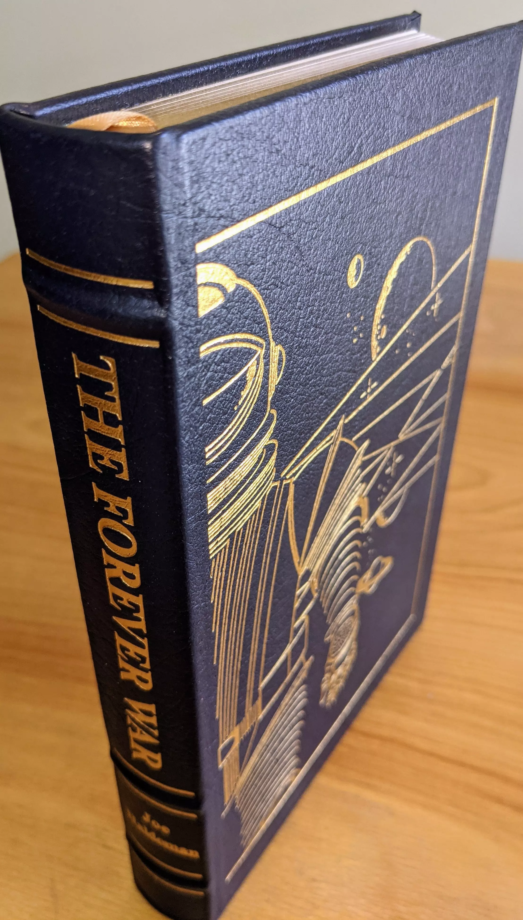 This is a collector's edition of The Forever War by Joe Haldeman, produced by The Easton Press as part of their Masterpieces of Science Fiction Collection. The book is bound in full dark navy blue leather, featuring raised hubbed spines and a cover accented in genuine 22kt gold
 - 
The front cover displays original artwork by Vincent DiFate, depicting a stylized astronaut in a helmet and suit, set against a space backdrop with planets and stars, all etched in 22k gold lines. A geometric 22k gold border frames the illustration, adding a refined touch. The spine is embossed with the title THE FOREVER WAR and the author's name in 22k gold, along with decorative horizontal bands
 - 
The volume is printed on archival-quality acid-neutral paper with 22k gold gilded edges, ensuring both durability and a luxurious appearance. Smyth sewing and concealed muslin joints contribute to the book's structural integrity and longevity. Inside the front cover is a pristine, unused bookplate, indicating its collector's status
 - 
This copy comes from a single owner's private collection, meticulously stored in dust-free bookcases in a dry climate, and remains in pristine, unread condition. The edition includes an introduction by Ben Bova, further enhancing its appeal for collectors. This stunning volume exemplifies high-quality craftsmanship and preservation, making it a standout piece for any science fiction enthusiast or collector seeking a flawless and visually striking edition