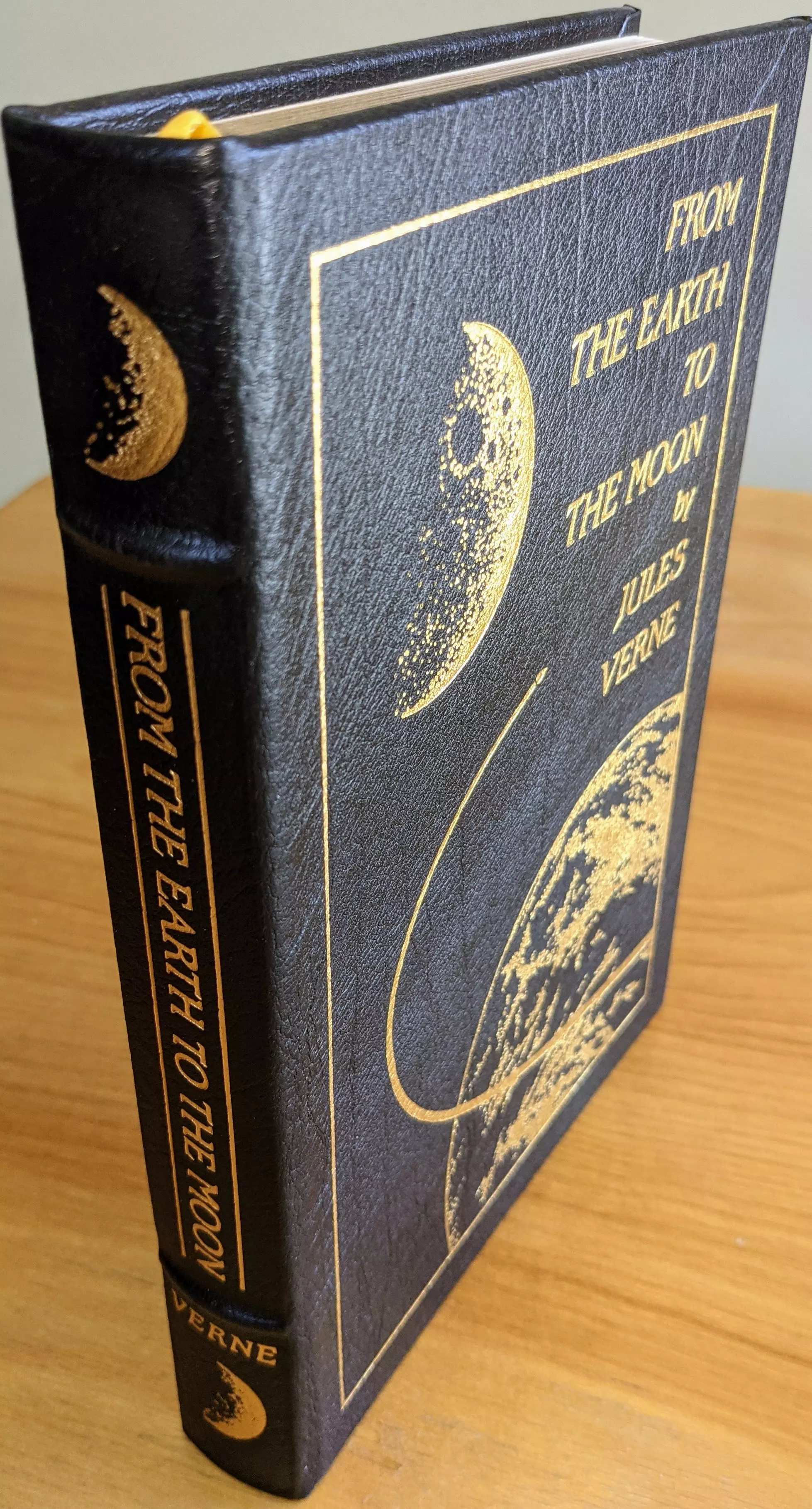 Pristine, leather-bound Collector Edition of From the Earth to the Moon by Jules Verne, published by The Easton Press in 1970 as part of the Masterpieces of Science Fiction Collection. Introduction by Jean Jules Verne
 - 
Bound in black leather, with elegant 22k gold-foil embossing. The cover has a gold illustration of the moon. Below it, the Earth is depicted in a similar golden style. A fine gold rectangular border frames the artwork. The title, From the Earth to the Moon, is embossed in bold gold lettering, positioned in the upper right quadrant of the cover, with Jules Verne beneath.

On the spine of the book, the title is prominently displayed in large, gold lettering, running horizontally along the spine. Below it, Verne, is inscribed in gold letters. At both the top and bottom of the spine, a crescent moon is embossed in gold. The spine also features raised hubs. 
 - 
The pages are printed on archival-quality, acid-neutral paper. The page edges are 22k gold gilded. Inside, the book includes a pristine bookplate and features an original frontispiece artwork by Robert Shore. The smyth-sewn binding ensures durability, while concealed muslin joints provide additional structural integrity
 - 
Meticulously preserved in a dust-free bookcase within a dry climate by a single private owner, this volume remains in pristine condition
 - 
A stunning and rare collectible for science fiction enthusiasts and fine book collectors alike