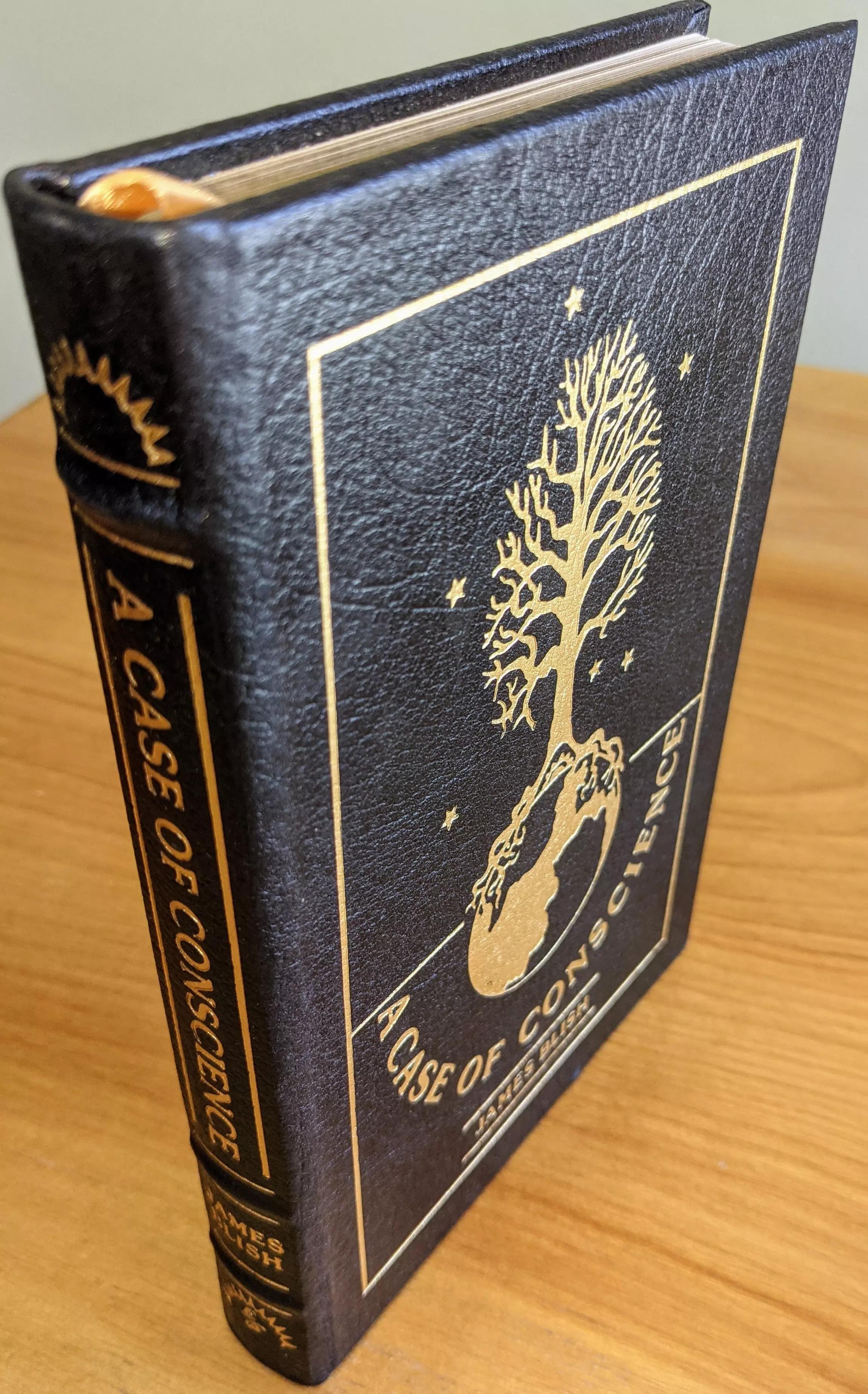 Exquisite Collector's Edition copy of: A Case of Conscience by James Blish from The Easton Press's Masterpieces of Science Fiction Collection, meticulously maintained by a single owner in a private collection, stored carefully in dust-free bookcases in a dry climate. Bound entirely in rich, black genuine leather, the volume is adorned with exquisite original artwork by Ellen Sull Farley, precisely accented in brilliant 22kt gold detailing
 - 
The front cover prominently features a striking illustration in 22kt gold foil depicting a detailed, symbolic image: a tree flourishing upward into branches, rooted deeply within a globe representing Earth, encapsulating the profound ethical and existential themes explored in the novel. Tiny golden stars surround the central design, adding elegance and cosmic depth to the artwork
 - 
The spine, luxuriously hubbed, displays the title A CASE OF CONSCIENCE and the author JAMES BLISH prominently in gleaming 22kt gold lettering, accompanied by intricate decorative motifs at both the top and bottom
 - 
Printed on archival-quality acid-neutral paper to ensure longevity, the pages feature gilded edges that complement the gold accents of the cover. Expert craftsmanship includes traditional smyth-sewn binding and concealed muslin joints, providing exceptional durability. The volume contains a pristine, untouched bookplate inside the front cover and features an introduction by Walter E. Meyers. Flawless, pristine, unread, and as new