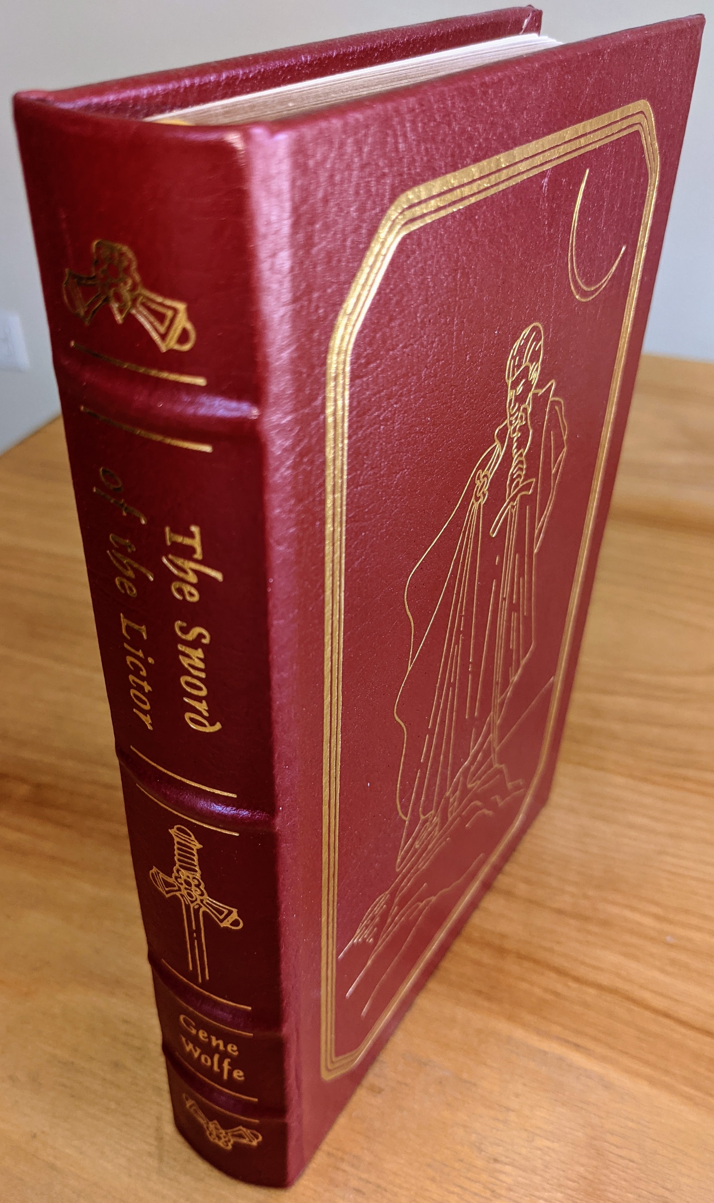 This luxurious volume of The Sword of the Lictor by Gene Wolfe is an exceptional collector's edition, published by The Easton Press as part of the Masterpieces of Science Fiction Collection. The book is bound fully in rich, deep burgundy leather, meticulously accented with brilliant 22kt gold detailing. The front cover features original artwork by artist Ron Miller, elegantly rendered in fine 22k golden lines. It depicts a cloaked figure holding a sword downward, standing on rocky terrain beneath a crescent moon, conveying a sense of dark mystery and quiet power
 - 
The spine, with its classic hubbed design, is ornately accented in 22k gold, displaying both title and author's name in crisp gold script alongside decorative motifs, including an intricately illustrated sword
 - 
The volume, produced in a limited run, includes an introduction by Gary K. Wolfe. It is printed on premium archival-quality, acid-neutral paper with 22k gold gilded edges, ensuring its preservation. Binding is robust, utilizing traditional smyth sewing with concealed muslin joints for strength and durability
 - 
Inside the front cover lies a pristine bookplate, untouched and ready for personalization. This copy, from a single-owner private collection, remains unread and has always been stored meticulously in dust-free bookcases within a dry climate. Its condition is impeccable, accurately described as pristine, as-new, and unread. A flawless specimen for a discerning collector