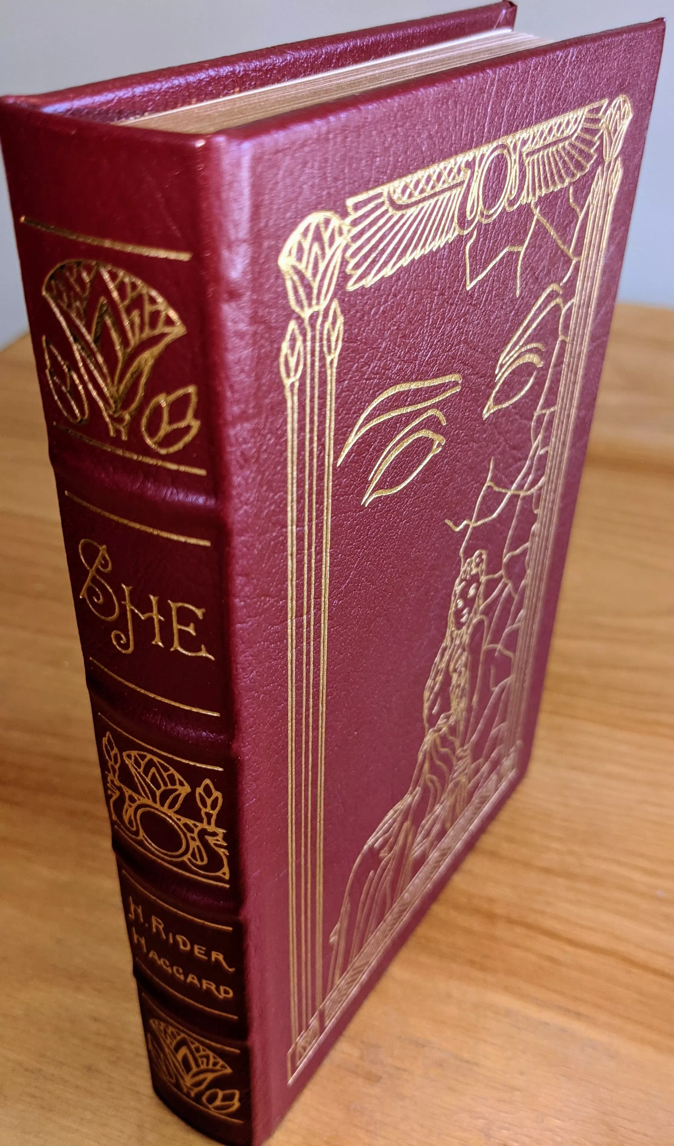 Stunning collector's edition of She by H. Rider Haggard, issued as part of the Masterpieces of Science Fiction series. Bound in full burgundy leather, accented in 22kt gold. The spine has hubbed bands and features 22k gold-stamped decorative elements and typography. The word SHE appears in elegant script at the top, followed by intricate ornamental flourishes. The author's name, H. Rider Haggard, is stamped in gold at the base of the spine
 - 
The front cover displays original artwork by Frank Kelly Freas, rendered entirely in 22k gold embossing. The illustration features the stylized face of a woman formed from sweeping contour lines, her eyes, eyebrows, and part of her nose are outlined with precision. To the right, a vertical crack disrupts the face, within which a smaller, full-body figure is visible, draped in long, flowing robes and standing in a mysterious, elevated pose. Vertical decorative lines frame the sides, topped with botanical motifs that resemble stylized buds or flames. The top of the frame includes a central circular symbol flanked by symmetrical wings or fanned forms
 - 
This edition includes an introduction by Everett F. Butler. It is printed on archival-quality paper with gilded page edges, Smyth sewing, and concealed muslin joints. The book includes a pristine bookplate on the inside front cover. Condition is Pristine / As New / Unread. Single-owner copy from a private collection, always stored in dust-free bookcases in a dry climate. Limited run