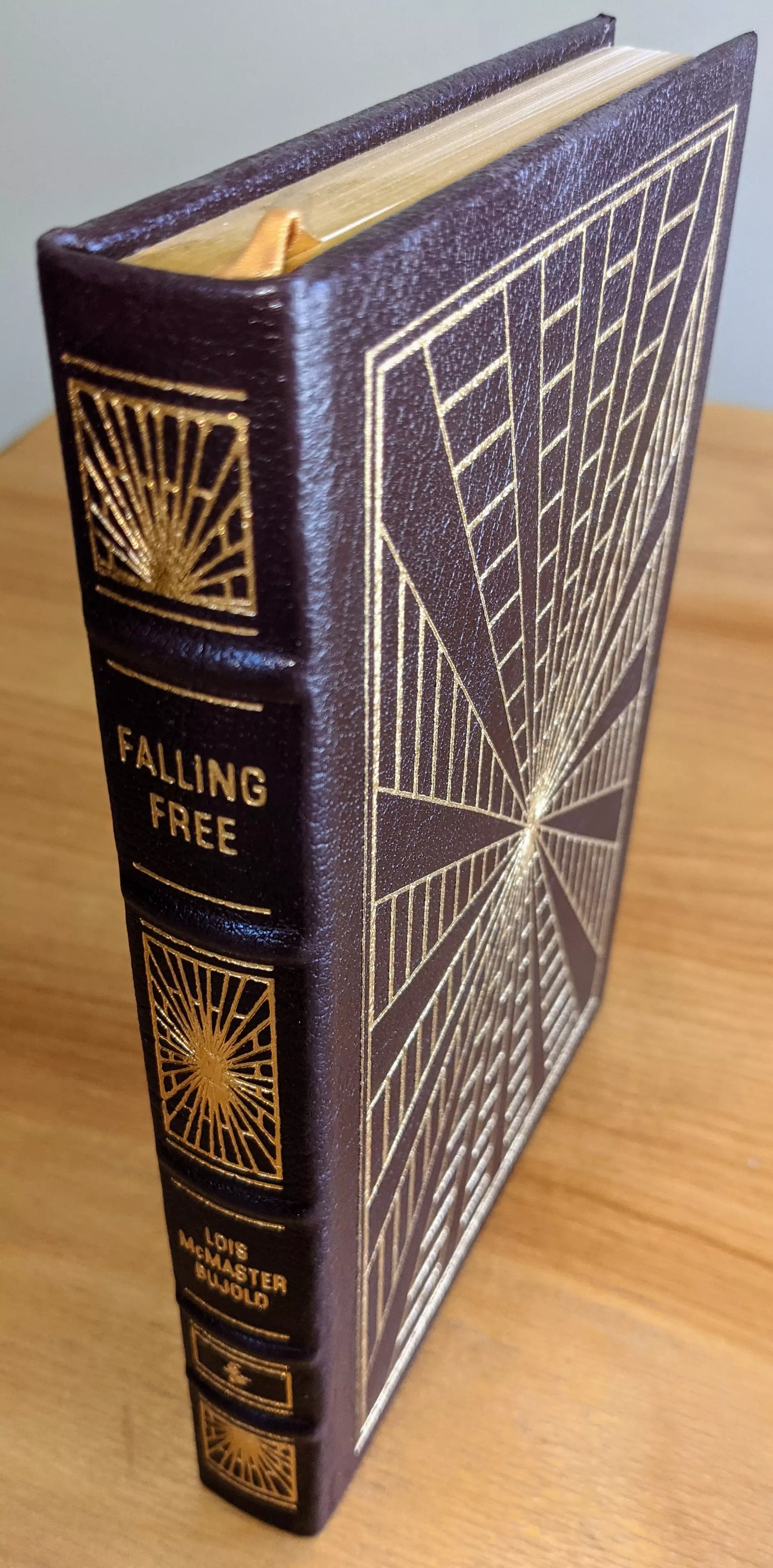 This is a deluxe, collector-grade edition of Falling Free bound in deep, rich brown leather with a smooth, refined texture and precise craftsmanship
 - 
The cover features an elaborate geometric sunburst style pattern, meticulously stamped in radiant 22k gold, with sharp, clean lines radiating outward in symmetrical angular segments 
 -
The spine is hubbed and equally ornate, with raised bands and gilt stamped panels, including the title, the author's name Lois McMaster Bujold, and matching decorative burst emblems
- 
The page edges are fully gilded, gleaming smoothly and evenly, and the volume includes a gold satin ribbon marker tucked neatly between pages
 - 
The binding appears tight and expertly sewn, reflecting archival-quality smyth sewing and reinforced internal joints
 - 
From The Easton Press Masterpieces of Science Fiction series, printed on acid-neutral archival paper. The bookplate inside the front cover is unused and pristine. Stored in dust-free cases in a dry setting to keep the leather, gilding, and paper immaculate. The artwork and design reference the publisher's tradition of fine, timeless presentation, though the specific artist is not credited
 - 
Overall, the volume remains in exceptional, flawless collectible state with bright gold accents, straight boards, sharp corners, and no visible wear presenting a striking, elegant science-fiction classic in premium form