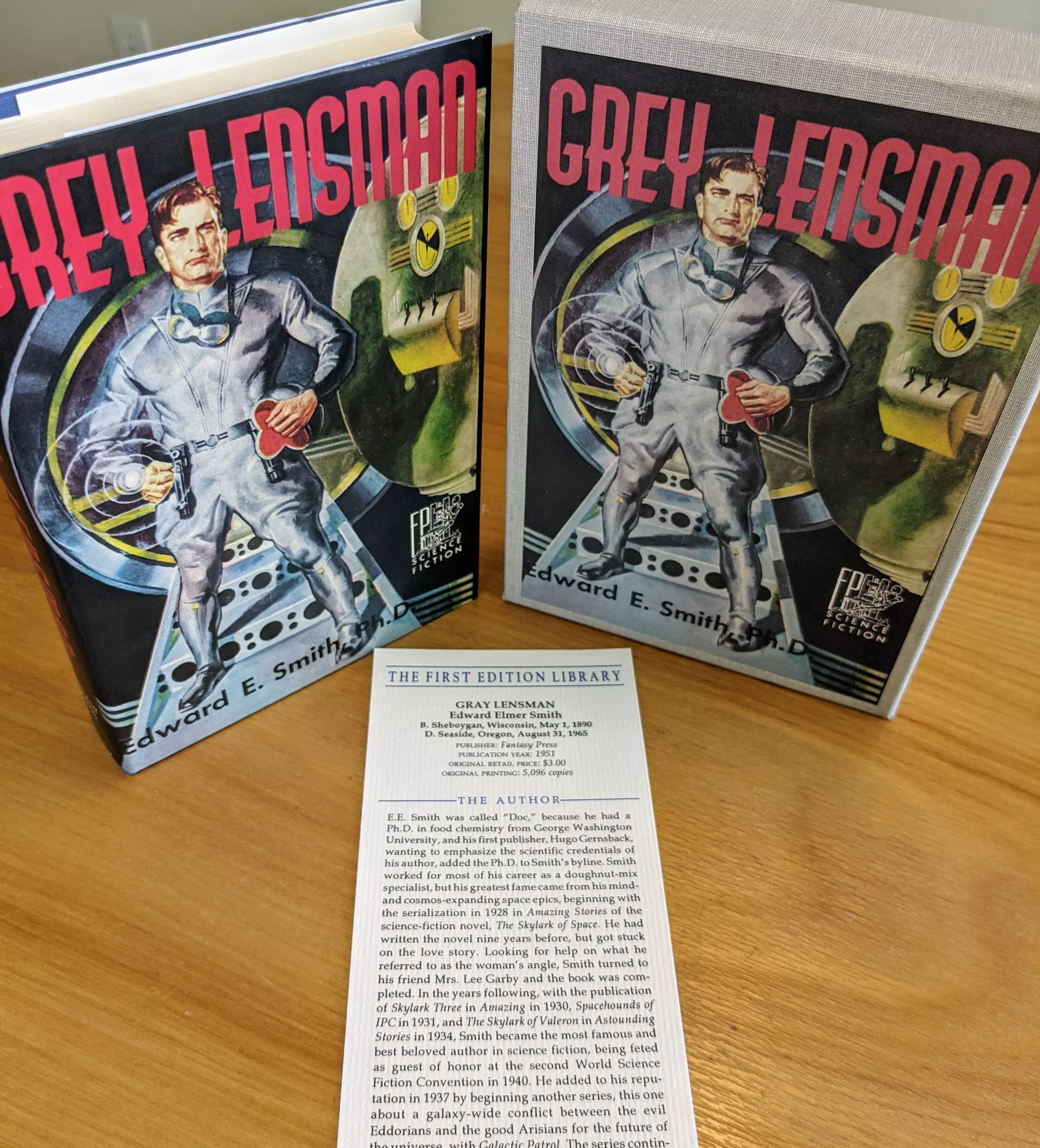 The cover of Grey Lensman by Edward E. Smith, Ph.D., reproduced here as a facsimile first edition by The First Edition Library, is a precise recreation of the 1951 Fantasy Press original. This copy, from a single-owner private collection, is unread, stored in dust-free bookcases in a dry climate, and preserved in pristine condition. It includes the original-style dust jacket, a matching cloth slipcase, and a publisher's bookmark with bibliographic details and historical context, making it both a collector's item and a reference edition
 - 
The artwork features the titular Grey Lensman standing confidently on a spacecraft deck. He wears a metallic-gray uniform and black boots. His right wrist displays the Lens—a glowing circular device emitting energy waves, identifying him as a member of the Galactic Patrol. In his left hand, he holds a red lined black helmet. His posture is assertive, facing forward with feet planted wide. Behind him is a large circular doorway flanked by two control consoles in green and yellow, filled with gauges and switches, suggesting a command station
 - 
The title GREY LENSMAN appears in large, red, block letters across the top. The author's name, Edward E. Smith, Ph.D., is printed at the bottom in white. This facsimile, part of a series of over 100 flawless reproductions issued by the First Edition Library in the 1980s-1990s, maintains the original typography, layout, and even typesetting quirks, making it both a collectible and scholarly reference