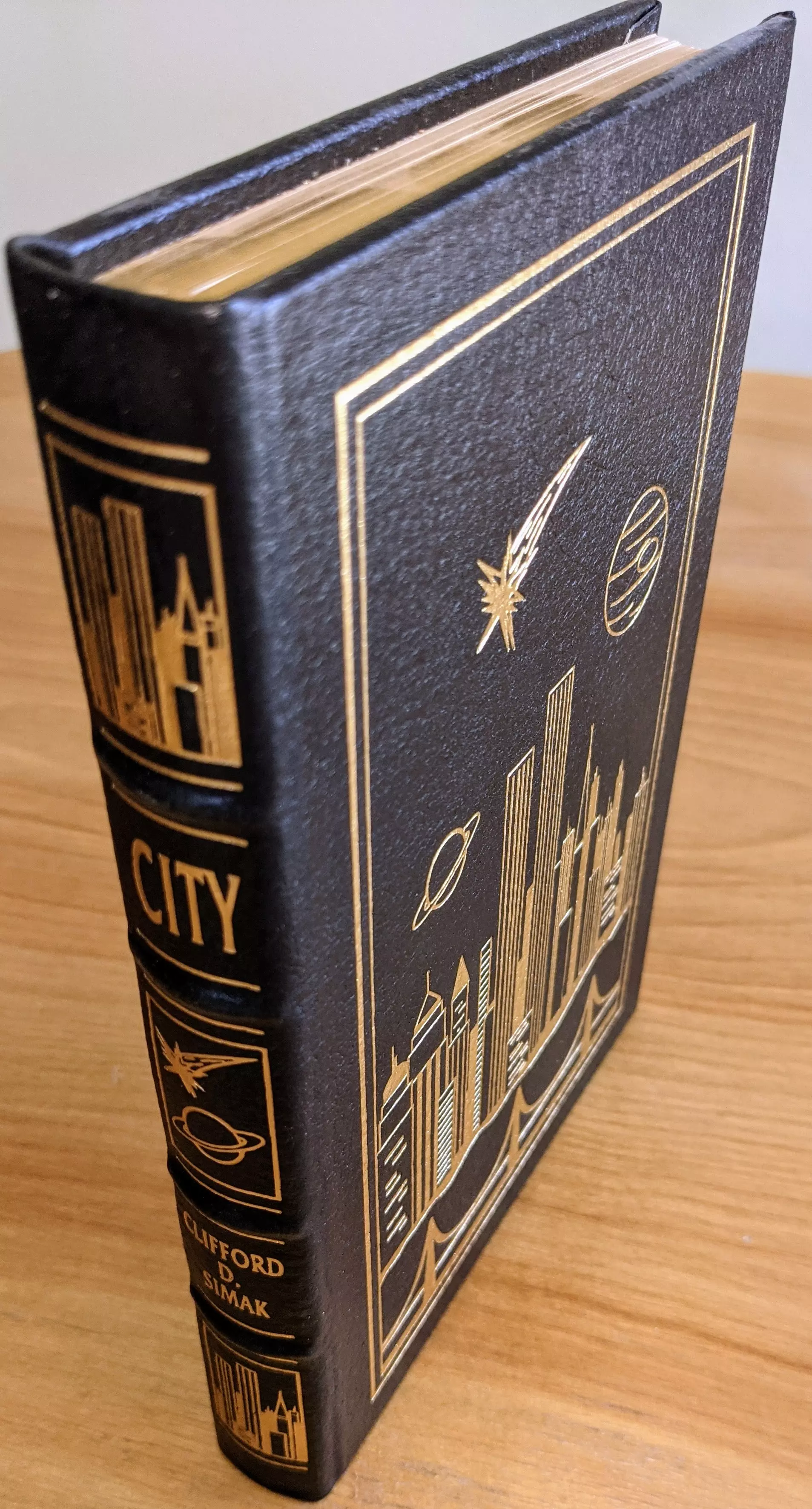 Pristine, unread (1995) collector's edition of Clifford D. Simak's City, from a single-owner private collection, stored in dust-free bookcases in a dry climate. This limited-run volume is part of the Masterpieces of Science Fiction Collection by The Easton Press
 - 
The book is bound in full black leather with hubbed spines, featuring original cover artwork by Pat Morrissey. The front and spine are accented in 22kt gold, with intricate, sharp gold-stamped illustrations: a stylized city skyline, planets, and a comet, all framed by double gold lines. The spine displays the title CITY and the author's name, CLIFFORD D. SIMAK, in large gold letters, along with additional gold-embossed cityscape and planetary motifs
 - 
The book is printed on archival-quality acid-neutral paper with 22k gold gilded edges, ensuring long-term preservation and a luxurious appearance. Smyth sewing and concealed muslin joints provide superior durability and allow the book to open flat without damage. Inside, a pristine bookplate is affixed to the front cover, untouched and ready for personalization
 - 
This edition includes a specially commissioned introduction by Robert Silverberg, adding scholarly context and collectible value. The overall condition is as new, with no signs of handling or wear, making it a flawless example for collectors. This volume exemplifies Easton Press's commitment to craftsmanship, combining fine materials, original art, and literary significance in a single, exceptional book