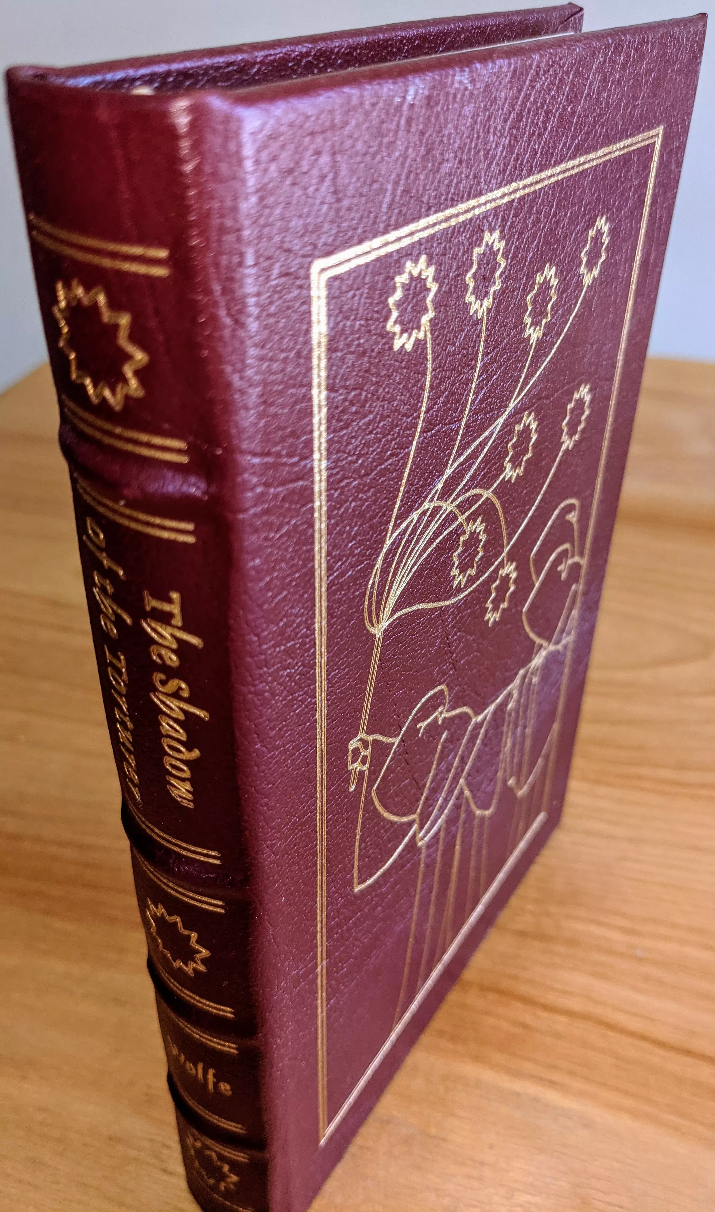 Stunning volume of The Shadow of the Torturer by Gene Wolfe is an exquisite collector's edition published by The Easton Press as part of the Masterpieces of Science Fiction Collection. Bound fully in luxurious burgundy leather, the cover features elegant original artwork by Michael Mariano, precisely accented in lustrous 22kt gold. The front displays an intricate scene outlined in 22k gold: a stylized, hooded figure holding a torture's flail, set within a clean rectangular frame. The spine is beautifully hubbed, divided into compartments separated by detailed 22k gold bands, displaying the title and author's surname in graceful 22k gold script
 - 
This limited-run volume is introduced by Tom Shippey and is expertly printed on premium acid-neutral archival paper designed to withstand aging. Its edges are flawlessly gilded, adding refinement and elegance. The craftsmanship includes traditional smyth sewing and concealed muslin joints, guaranteeing lasting durability and stability for collectors
 - 
Included is a pristine bookplate carefully placed on the inside front cover, untouched and ready for personalization. From a single-owner private collection, this book has remained unread since publication and always stored in dust-free bookcases within a carefully maintained dry climate. The volume's condition remains pristine, truly as new, making this rare, luxurious leather-bound book a striking and impeccable addition to any fine science fiction/fantasy collection