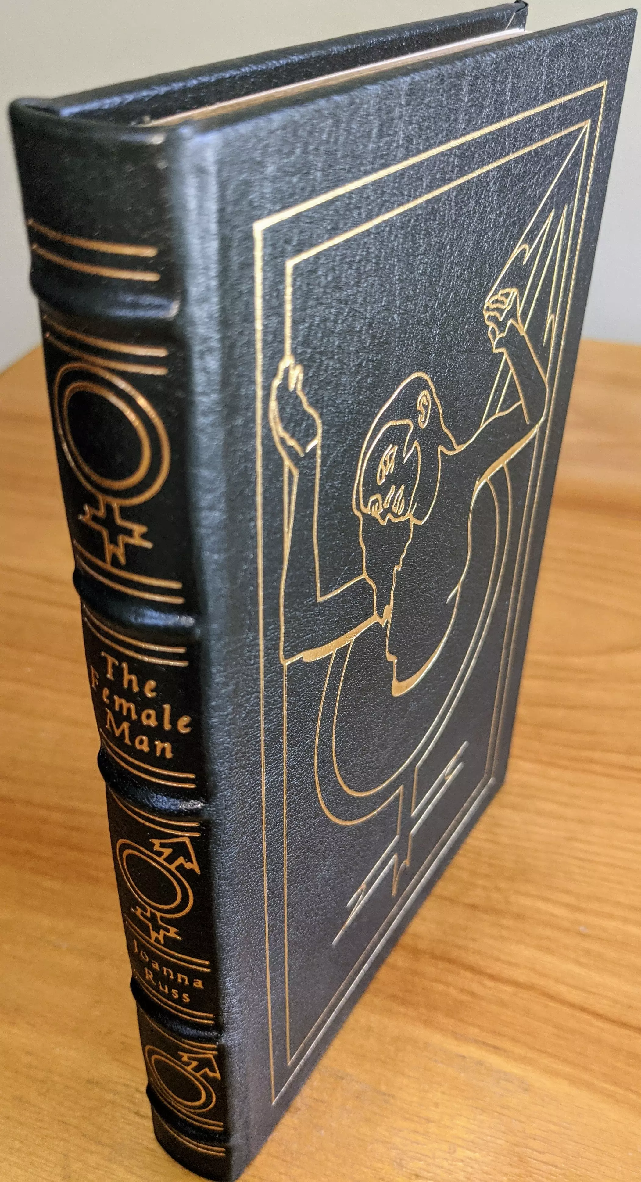 A stunning and rare collectible collector's edition of The Female Man by Joanna Russ, published (1994) as part of The Masterpieces of Science Fiction Collection by The Easton Press. Introduction by Sarah Lefanu, and original artwork by Clee Richeson
- 
Bound in full dark olive green leather, giving it a rich, elegant appearance. The spine features raised bands (hubbed spine) and is accented with 22kt gold, displaying the title, author's name, and gender symbols in striking gold detailing
 - 
The cover showcases a bold, gold-embossed minimalist illustration depicting a stylized figure emerging from a circular portal. The book includes a pristine bookplate on the inside front cover
 - 
The archival-quality, acid-neutral pages have gold gilded edges, enhancing its luxurious aesthetic, while smyth-sewn binding and concealed muslin joints ensure durability. This exceptional edition is a true collector's treasure and a striking presentation of Russ's groundbreaking feminist science fiction novel
 -  
This single-owner volume has been carefully stored in dust-free bookcases within a dry climate, preserving its pristine condition

