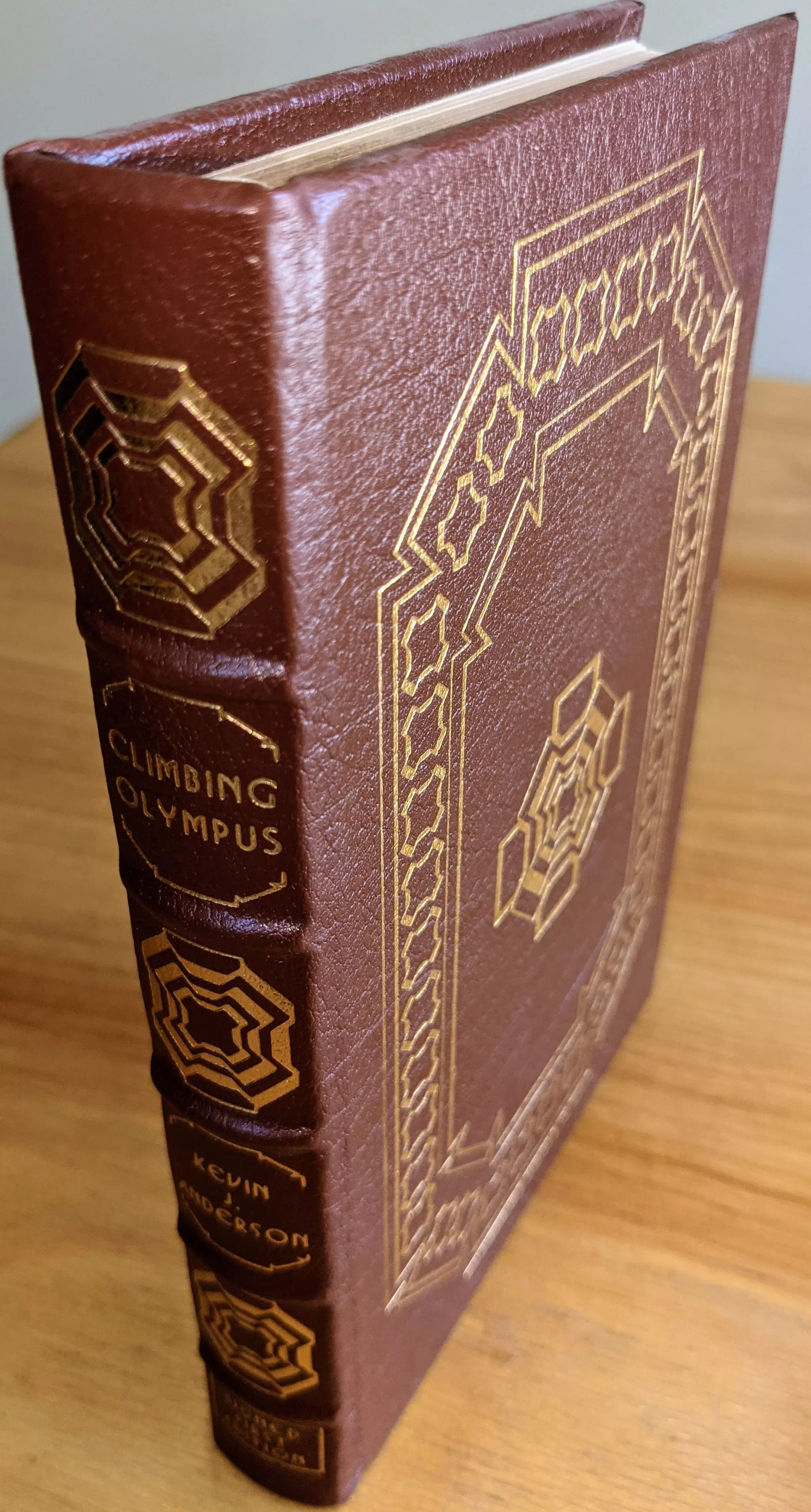 Stunning brown leather-bound first edition of Climbing Olympus by Kevin J. Anderson, part of The Signed First Editions of Science Fiction Collection by The Easton Press. The cover and spine are adorned with intricate gold-embossed geometric designs that exude elegance and sophistication. The title Climbing Olympus and the author's name, Kevin J. Anderson, are prominently displayed on the spine
 - 
This SIGNED collector's edition is bound in full leather with raised hubbed spines, a hallmark of Easton Press craftsmanship. The volume is accented in 22kt gold, printed on archival acid-neutral paper with 22k gold gilded edges. The binding features smyth sewing and concealed muslin joints, providing superior durability and a refined finish
 - 
Stored in pristine condition, this book is part of a single-owner private collection, carefully kept unread in dust-free bookcases within a dry climate to maintain its immaculate state. A pristine bookplate is included on the inside of the front cover, adding a personal touch to its collectibility. This limited-run edition is signed by Kevin J. Anderson, and introduced by James Gunn further increasing its value for collectors
 - 
Additionally, the book features stunning original artwork by Jeff Fisher, which perfectly complements Anderson's narrative. This extraordinary edition combines masterful craftsmanship, artistic brilliance, and literary significance, making it a prized addition for collectors and science fiction enthusiasts alike