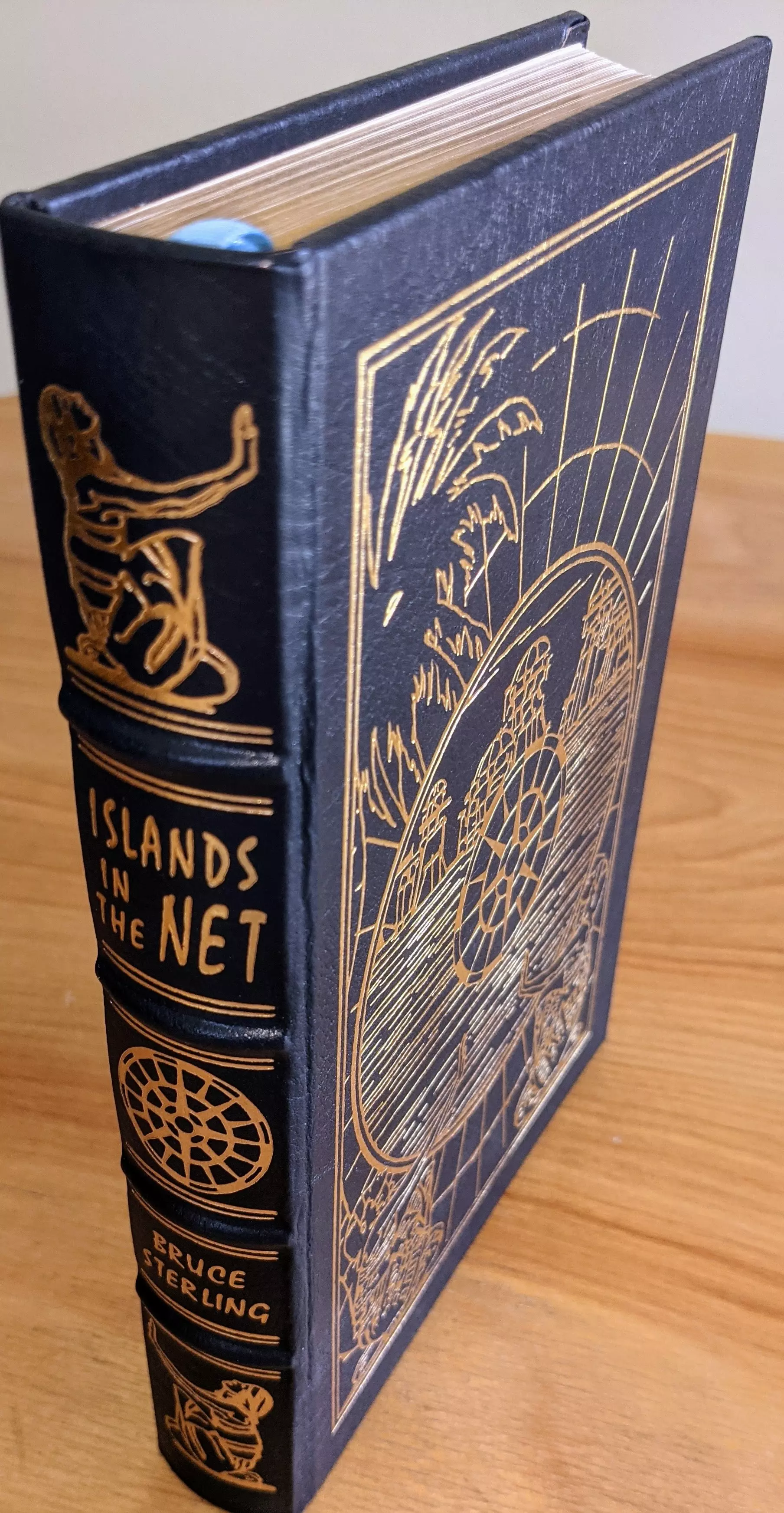 Stunning Easton Press limited collector's edition of Islands in the Net by Bruce Sterling from the prestigious Masterpieces of Science Fiction series. This edition is bound in full genuine leather, dyed a deep dark navy blue (almost black), with elaborate 22kt gold embossing on both cover and spine. The front cover features original artwork by Kent Bash, rendered in 22k gold leaf, with a network-like schematic surrounding an island paradise, encapsulated in a stylized grid structure. Introduction by James Gunn
 - 
The spine is hubbed and hand-tooled, featuring a geometric satellite-like emblem and two intricate vignettes of seated human figures using terminals—evoking both Sterling's cyberpunk roots and the narrative's geopolitical techno-surveillance themes. The title and author's name are boldly stamped in 22k gold, perfectly aligned with the edition's archival standards
 - 
Printed on acid-neutral archival paper, the pages are 22k gold gilded on all edges, giving the book a brilliant reflective sheen. Smyth sewing and concealed muslin joints ensure a lifetime of structural integrity. This copy is unread, from a single-owner private collection, and has been stored exclusively in dust-free, climate-controlled bookcases—preserved in pristine, as-new condition. The bookplate inside the front cover remains untouched. A true artifact for collectors, showcasing craftsmanship and literary significance in equal measure